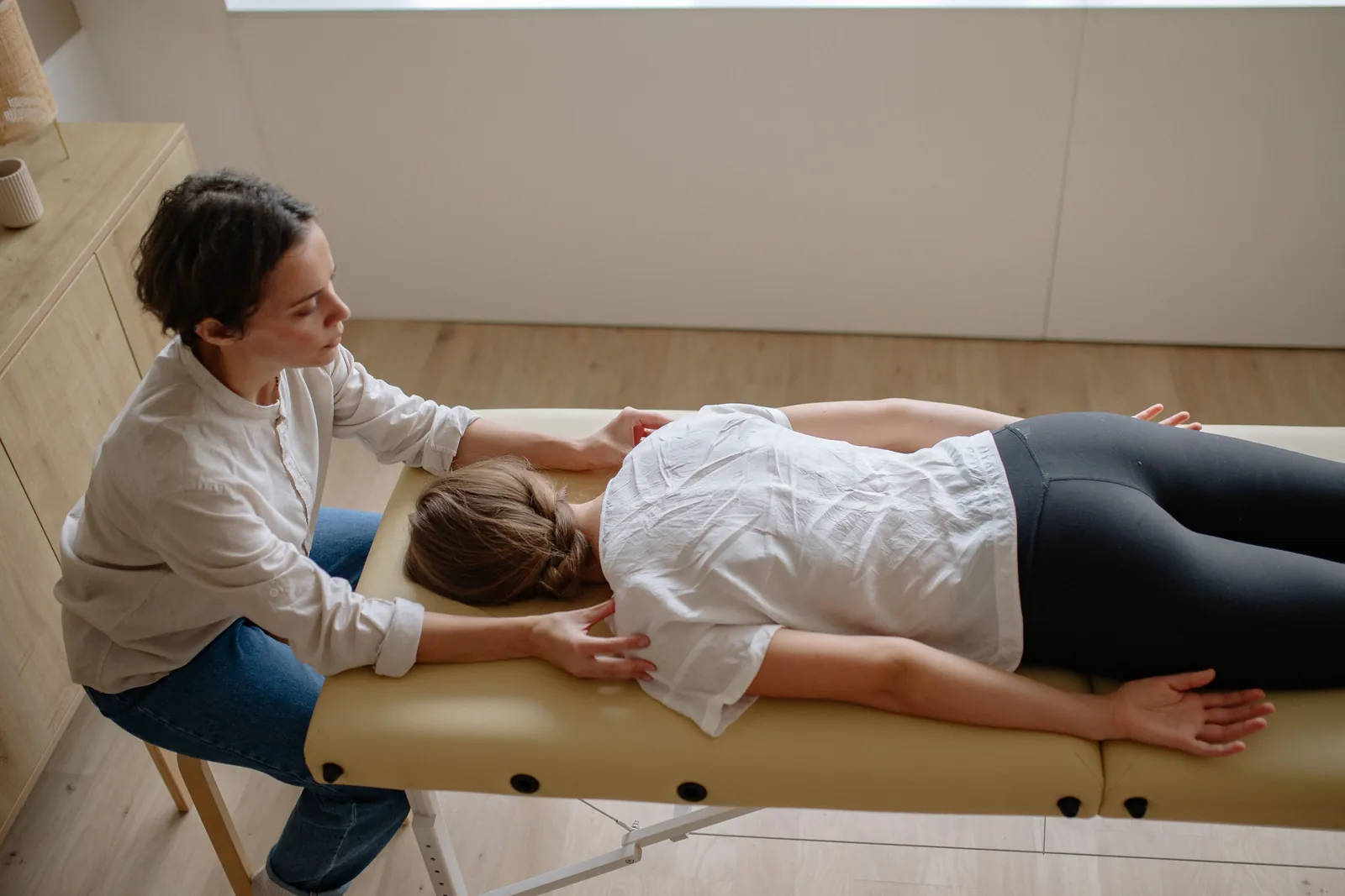 Practitioner's hands on the upper back of a woman lying face down on a treatment table
